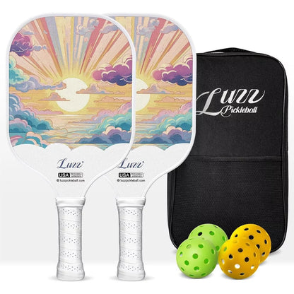 Two paddles arranged front and back, featuring sunrise-through-clouds artwork on the paddle faces and white handles. A black paddle bag, two indoor balls, and two outdoor balls are placed on the right side of the paddles.