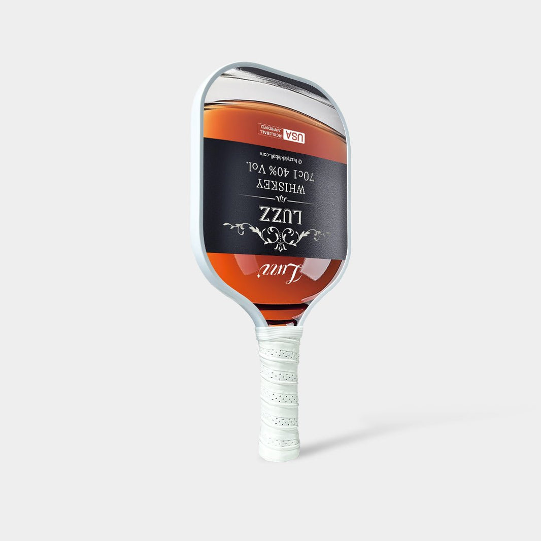 Luzz Whiskey Original Paddle standing upright at an angled front view, featuring a liquor bottle-shaped face with a Luzz logo in the center, USAPA certification logo at the top, white edge, and white handle.