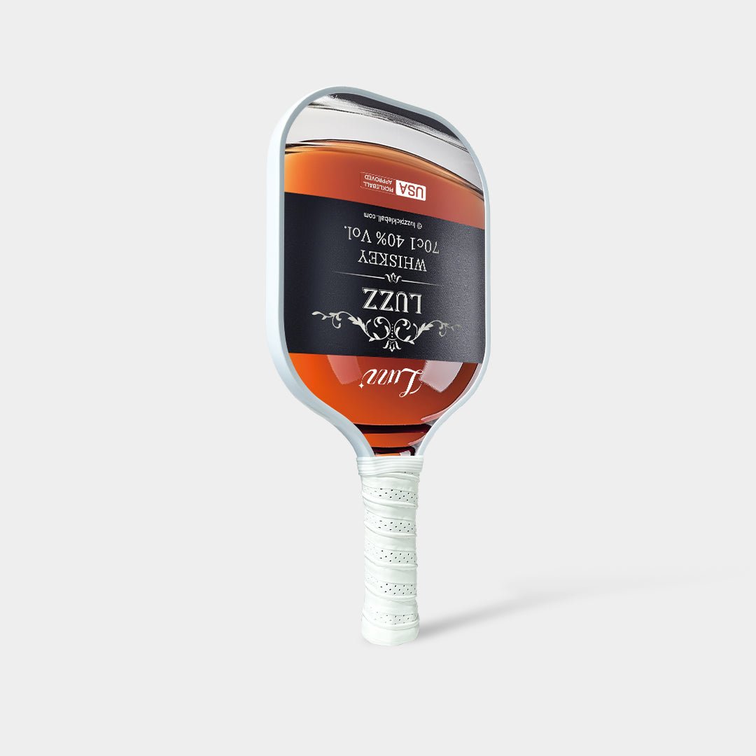 Luzz Whiskey Original Paddle standing upright at an angled front view, featuring a liquor bottle-shaped face with a Luzz logo in the center, USAPA certification logo at the top, white edge, and white handle.